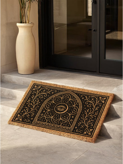 A rectangular, natural brown coir doormat with a non-slip black vinyl backing, viewed from directly above. The mat is made of textured coconut fiber. The design features intricate black line art in a Victorian Gothic and Art Nouveau style. In the center is an All Seeing Eye, or Eye of Providence, enclosed within a radiant starburst and a mandala-like floral pattern. This central motif is set inside an arched frame. The corners and sides are adorned with symmetrical, flowing botanical vines.